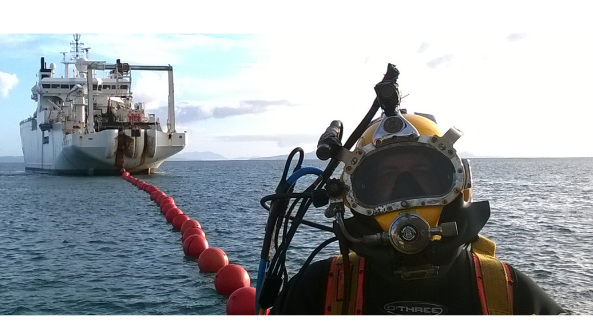 Diver in full helmet gear near a cable-laying ship deploying floating subsea cable markers.